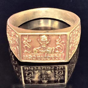 Op de afbeelding: Een goudkleurige ring met een rechthoekige voorkant met een reliëfbeeld van een figuur en tekst. De band van de ring is glad, terwijl de voorkant ingewikkelde details heeft. De ring wordt gepresenteerd op een reflecterend oppervlak.
