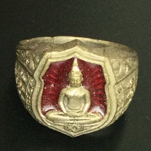 Op de afbeelding: Een goudkleurige ring met een zittende Boeddha-figuur in een rode, schildvormige setting. De ring heeft sierlijke details rond de randen en een donkere achtergrond.