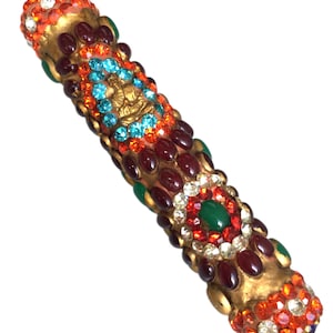 Puede incluir: Un brazalete dorado adornado con piedras preciosas rojas, verdes y azules. El brazalete está cubierto de pequeños diamantes de imitación transparentes y presenta una pequeña figura de Buda dorada en el centro.
