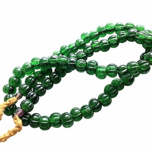 Puede incluir: Un collar de cuentas de vidrio verde oscuro y acanaladas. Las cuentas están ensartadas, formando un collar o elemento decorativo. Un cordón trenzado de color beige está unido al extremo del collar.