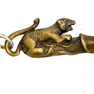 Può includere: Un portachiavi vintage in ottone con un'immagine di una tigre su una forma stilizzata. La tigre è dettagliata con linee incise e il ciondolo è attaccato a una catena color argento. Il design ha un'estetica antica.