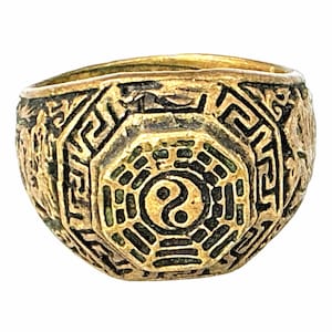 Op de afbeelding: Een goudkleurige metalen ring met een centraal achthoekig ontwerp met een yin-yang symbool omringd door zwart ingevulde trigrampatronen. De ring is breed en getextureerd met ingewikkelde geometrische Griekse sleutelmotieven en reliëfs aan de zijkanten.