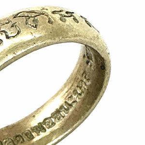 Op de afbeelding: Een zilverkleurige metalen ring met een getextureerd, verweerd oppervlak met in reliëf gegraveerde schrifttekens langs de buitenste band die doorlopen langs de binnenomtrek van de cirkelvormige metalen ring.