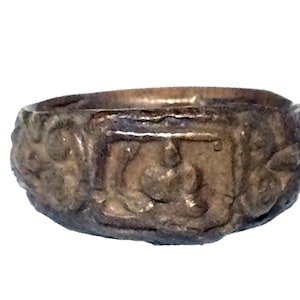 Puede incluir: Un anillo de plata con un diseño detallado. El anillo presenta un centro de forma cuadrada con una imagen en relieve de una persona. El anillo está rodeado por un borde decorativo.