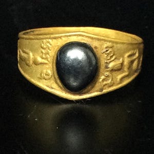 Puede incluir: Un anillo de oro con una piedra oscura de forma ovalada en el centro. La banda del anillo presenta diseños en relieve. El anillo está sobre un fondo oscuro, resaltando su brillo metálico y la superficie pulida de la piedra.