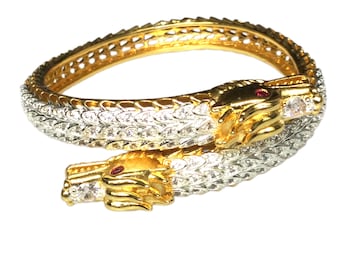 May include: A gold and silver-toned bracelet in the shape of a snake. The bracelet features two snake heads with red gemstone eyes and is encrusted with clear crystals. The bracelet is a unique piece of jewelry.