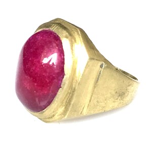 May include: A gold-toned ring featuring a large, oval-shaped, deep pink gemstone. The ring's band has a geometric design with a slightly textured surface. The gemstone is smooth and polished, reflecting light.