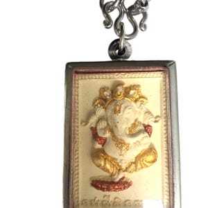 May include: A silver chain necklace with a rectangular pendant featuring a white and gold Ganesha statue. The pendant has a clear protective cover and a red and gold border.