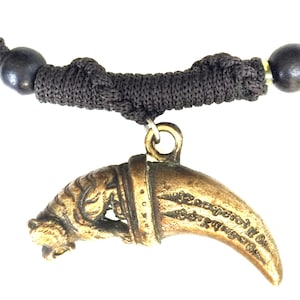 Puede incluir: Un colgante de color dorado en forma de diente de tigre con una pequeña figura de tigre en el lateral. El colgante está unido a un collar de cordón negro con cuentas de madera negras.