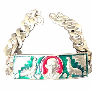 May include: Silver-toned bracelet with a rectangular centerpiece featuring a portrait in a red circle, flanked by green and white bird designs. The bracelet has a thick chain link design.