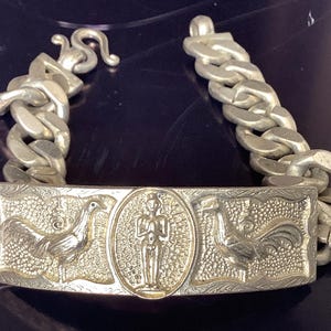 May include: Silver-toned bracelet with a rectangular centerpiece and a chain link design. The centerpiece features embossed images of roosters and a central figure. The bracelet is approximately 8 inches long.