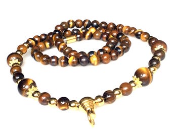 May include: A necklace featuring tiger eye beads in shades of brown and gold. The necklace has a gold-colored clasp and decorative elements. The beads vary in size, with larger beads interspersed among smaller ones. The necklace is displayed against a white background.