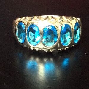 Op de afbeelding: Een goudkleurige ring met vijf ovale, helderblauwe edelstenen. De band van de ring heeft een textuurontwerp en de edelstenen zijn dicht bij elkaar geplaatst, waardoor een doorlopende lijn van kleur ontstaat. De ring wordt op een donkere ondergrond gepresenteerd.