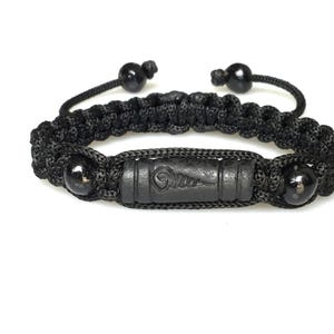 May include: Black braided bracelet featuring a central rectangular bead and two spherical beads. The bracelet is adjustable with black cord and has a carved design on the central bead. The bracelet is set against a white background.