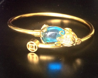 Puede incluir: Un brazalete de oro con una piedra preciosa azul y un dije de rana de oro. El brazalete tiene un símbolo chino en el lateral.