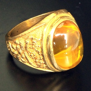 May include: A gold ring with an oval-shaped amber gemstone. The ring has a detailed design on the band.