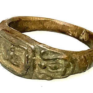 Puede incluir: Anillo antiguo de bronce con una cara rectangular y detalles ornamentados. El anillo presenta un aspecto envejecido, mostrando intrincados patrones y diseños. La banda es gruesa, lo que sugiere un origen histórico o vintage.