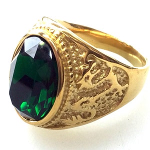 Può includere: Un anello d'oro con una grande pietra preziosa verde di forma ovale. L'anello presenta un design dettagliato sulla fascia, che rappresenta un drago.