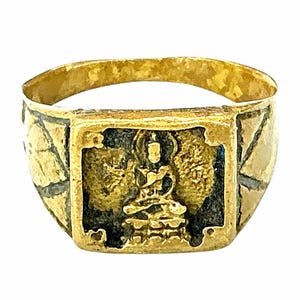 Op de afbeelding: Een goudkleurige metalen ring met een rechthoekig voorvlak met een verhoogde, zittende figuur tegen een donkere, verzonken achtergrond. De band vertoont een gestructureerd, geometrisch patroon met donkere lijnen die gouden driehoekige delen scheiden.