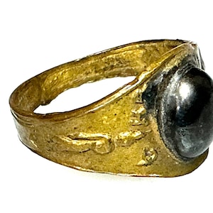 Puede incluir: Un anillo dorado con una piedra grande y oscura de forma ovalada. La banda del anillo tiene una superficie texturizada con detalles grabados. La piedra está engastada en un bisel plateado, creando un contraste con el oro.