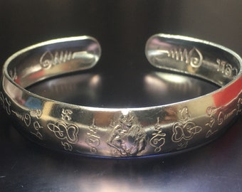 Puede incluir: Pulsera de plata con grabados intrincados y una imagen central de una deidad. La pulsera está adornada con símbolos y diseños repetidos.