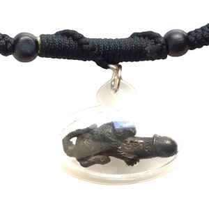 Può includere: Una collana con cordoncino nero con perline nere e un ciondolo ovale trasparente che contiene una piccola figurina in metallo nero.