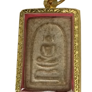 Könnte beinhalten: Ein goldfarbener Anhänger mit einem rechteckigen Rahmen und einer kleinen, verzierten Schlaufe oben. Der Rahmen hat einen roten Rand und ein weißes Innere mit einem erhabenen Bild eines sitzenden Buddha.