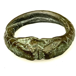 Puede incluir: Un anillo de bronce con un diseño intrincado y texturizado. El anillo tiene una apariencia oscura y envejecida con toques de pátina verde, lo que sugiere antigüedad. La banda tiene aproximadamente 2.5 cm de diámetro, con tallas detalladas alrededor de la superficie exterior.