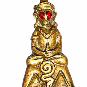 Pode incluir: Uma estatueta de metal cor de bronze de uma figura sentada com um toucado pontiagudo e duas pedras circulares vermelho-brilhante incrustadas nas órbitas oculares. A figura está sentada de pernas cruzadas com um design em espiral na base.