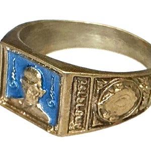Op de afbeelding: Een goudkleurige metalen ring met een rechthoekig voorvlak voorzien van een blauwe emaille inleg met een profielportret. De zijkant van de ring toont de in reliëf aangebrachte tekst "B.S.B.L.B.Q." en een rond decoratief embleem met een centraal punt.