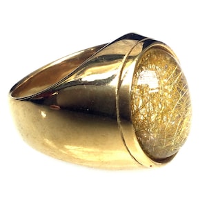 May include: A gold ring with a large, oval-shaped, clear stone with gold inclusions. The stone is set in a gold band with a smooth, rounded surface.
