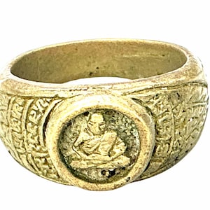 Op de afbeelding: Een metalen ring met een verweerde goudkleurige afwerking met een cirkelvormig centraal medaillon dat een zittende figuur in een meditatieve houding afbeeldt. De band is breed en versierd met ingewikkelde reliëfpatronen en tekst aan de zijkanten.