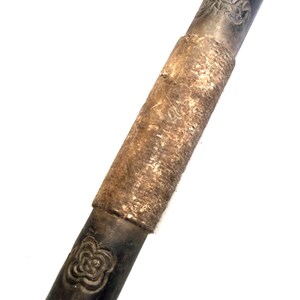 Può includere: Un oggetto cilindrico in pietra di colore grigio scuro, con una superficie ruvida di consistenza marrone. L'oggetto presenta un disegno decorativo a ciascuna estremità.