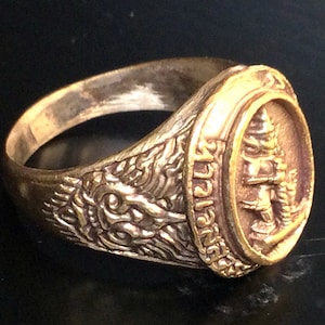 Puede incluir: Un anillo de color dorado con un diseño ovalado que presenta una imagen detallada de una figura de pie y escritura tailandesa. La banda del anillo está decorada con un diseño texturizado.