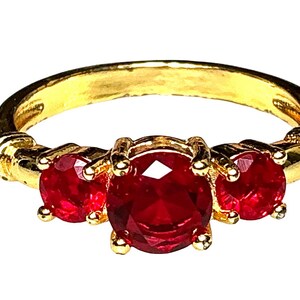 Puede incluir: Un anillo de oro con tres piedras rojas redondas y facetadas dispuestas en fila horizontal. La piedra central es más grande que las dos piedras laterales. La banda de oro tiene un acabado liso y pulido con detalles acanalados decorativos.