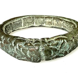 Op de afbeelding: Een antieke bronzen ring met een gedetailleerd ontwerp. De ring heeft een getextureerd oppervlak en een gespleten ontwerp met twee dierlijke figuren die tegenover elkaar staan. De ring heeft een verweerde, verouderde uitstraling, met hints van groene patina.