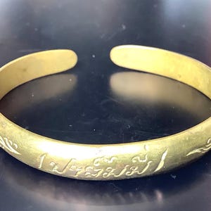 May include: A gold-colored open cuff bracelet. The bracelet has etched designs and text. The open end allows for easy wear. The bracelet is a simple, yet elegant piece of jewelry.