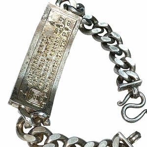 May include: A silver-toned metal chain bracelet featuring a rectangular pendant with engraved Thai script and a square geometric symbol. The bracelet is constructed with thick, interlocking oval links and includes a hook-style clasp closure.