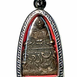 Peut inclure: Un pendentif en forme d'arche pointue présentant une figurine métallique sombre en position assise, enchâssée dans un cadre argenté avec une bordure intérieure rouge. Le pendentif est suspendu à une boucle métallique argentée.