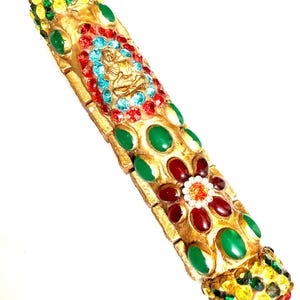 May include: A gold-colored decorative item adorned with colorful gemstones. The rectangular object features a central design with a seated figure surrounded by red and blue gems. Green, red, and yellow gems are also incorporated into the design.