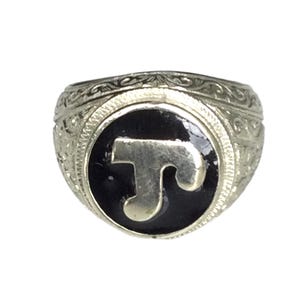 Op de afbeelding: Een zilveren ring met een sierlijk ontwerp en een zwart geëmailleerd centrum met een gouden letter "T".