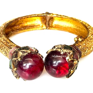 Peut inclure: Un bracelet doré articulé avec des détails ornés. Le bracelet présente deux grandes pierres précieuses rondes et rouge foncé au niveau du fermoir. Les extrémités du bracelet présentent des motifs texturés complexes. Le logo Jubilee Store est visible.