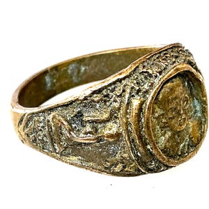 May include: A bronze-toned metal ring featuring a weathered, textured surface with a raised relief design. The centerpiece displays a circular medallion with a human face, while the band is decorated with an embossed figure in a reclining pose.