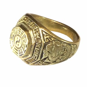 Op de afbeelding: Een goudkleurige ring met een achthoekig oppervlak met een Yin Yang-symbool. De band van de ring heeft gedetailleerde gravures, waaronder een feniksontwerp aan één kant. De ring is gemaakt van glanzend metaal.