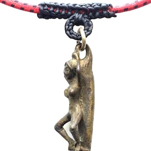 Può includere: Un ciondolo in bronzo con una figura femminile in piedi su una tavola da surf. Il ciondolo è attaccato a un cordino intrecciato rosso e nero.