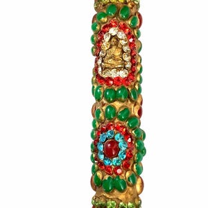 May include: A decorative, cylindrical object with a gold base, adorned with green, red, and clear gemstones. A central section features a gold Buddha figure surrounded by red and clear gems. The top and bottom are also embellished with colorful gems.