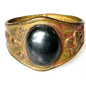 Puede incluir: Un anillo de estilo vintage con una piedra grande, ovalada, de color gris oscuro engastada en una banda dorada. La banda presenta patrones florales en relieve y detalles en rojo, creando una estética antigua única.