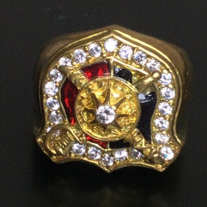 Puede incluir: Un anillo de oro con un diseño de esmalte negro y rojo, rodeado de piedras transparentes. El centro del diseño presenta una estrella con una piedra transparente grande.
