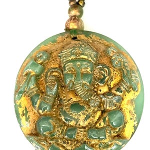 May include: Green and gold pendant necklace featuring a detailed relief of a deity. The pendant has a circular shape with a textured surface and a beaded necklace. The text "Jubilee Store" is visible on the pendant.
