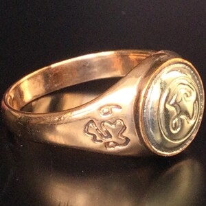 May include: A gold signet ring with a circular design featuring a stylized animal head. The ring has a smooth band and a raised design on the top.